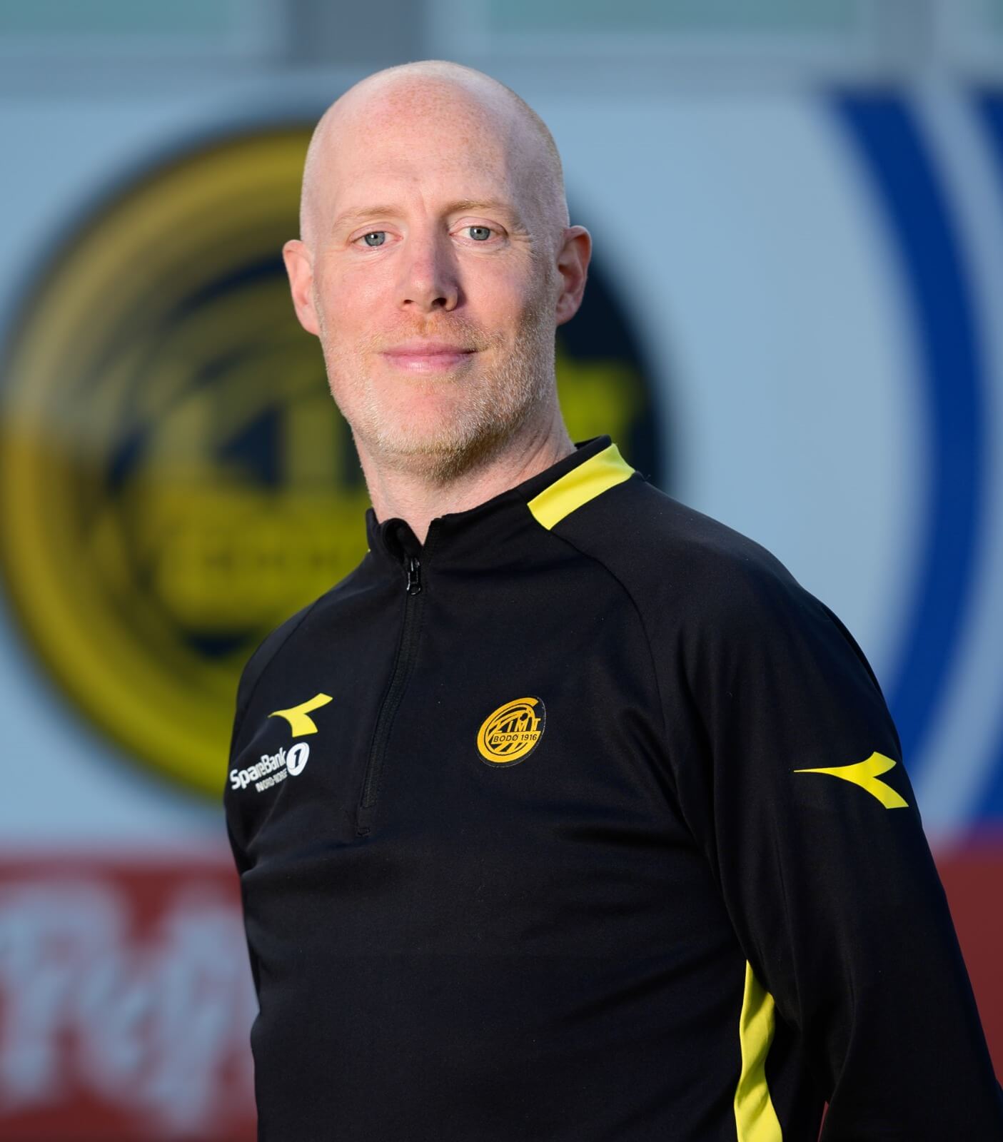 Gregg Broughton sonríe en el Bodo Glimt Field, vistiendo los colores negro y amarillo del equipo.